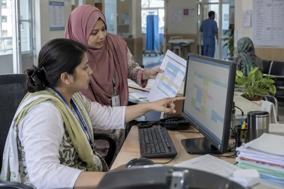 At a hospital scheduling desk, two administrative coordinators align an unmarked weekly consultant roster with appointment blocks on a monitor.