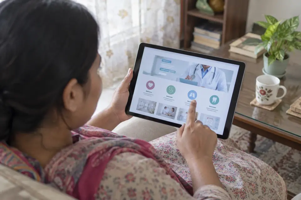 A patient at home views a clinic website's departments and contact paths on a front-facing tablet, with the screen content kept generic and text unreadable.