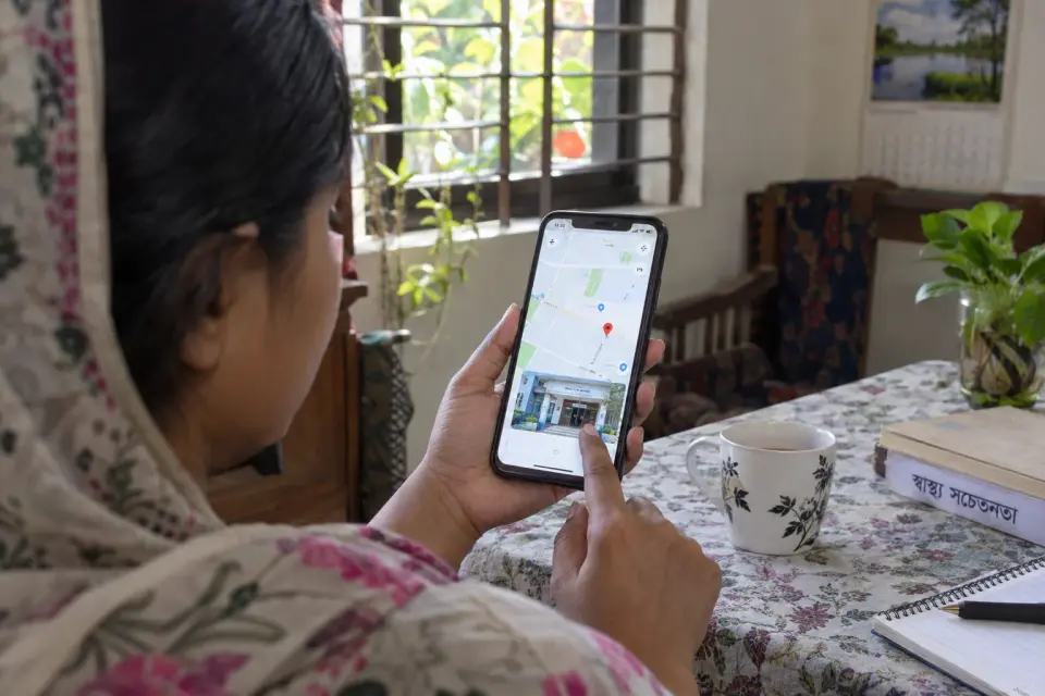 At home in Bangladesh, a patient checks the doctor's actual chamber area on a front-facing phone map while planning a visit; no invented address.